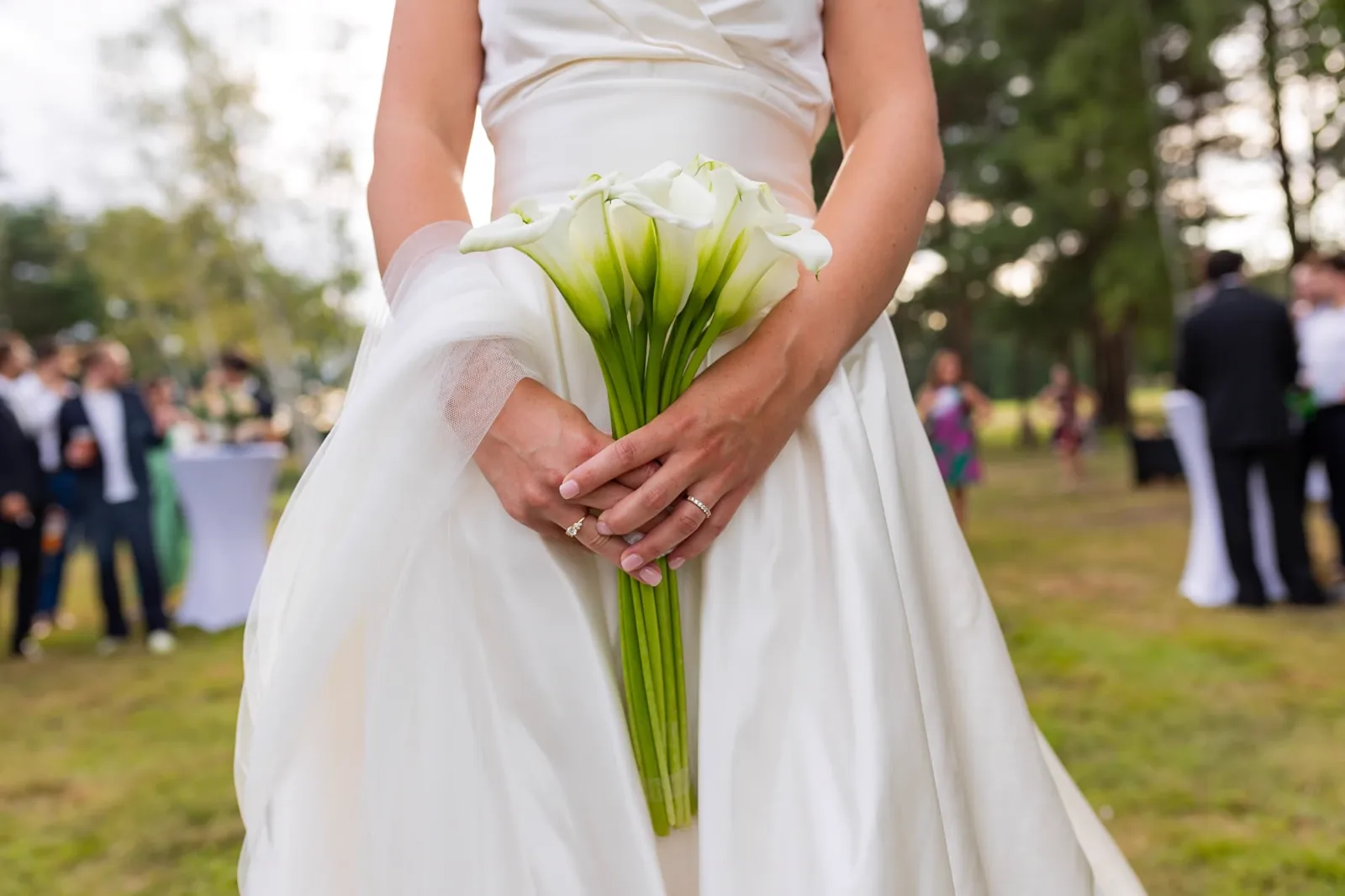 Mariée tenant son bouquet d’arums blancs lors du vin d’honneur, invités en arrière-plan