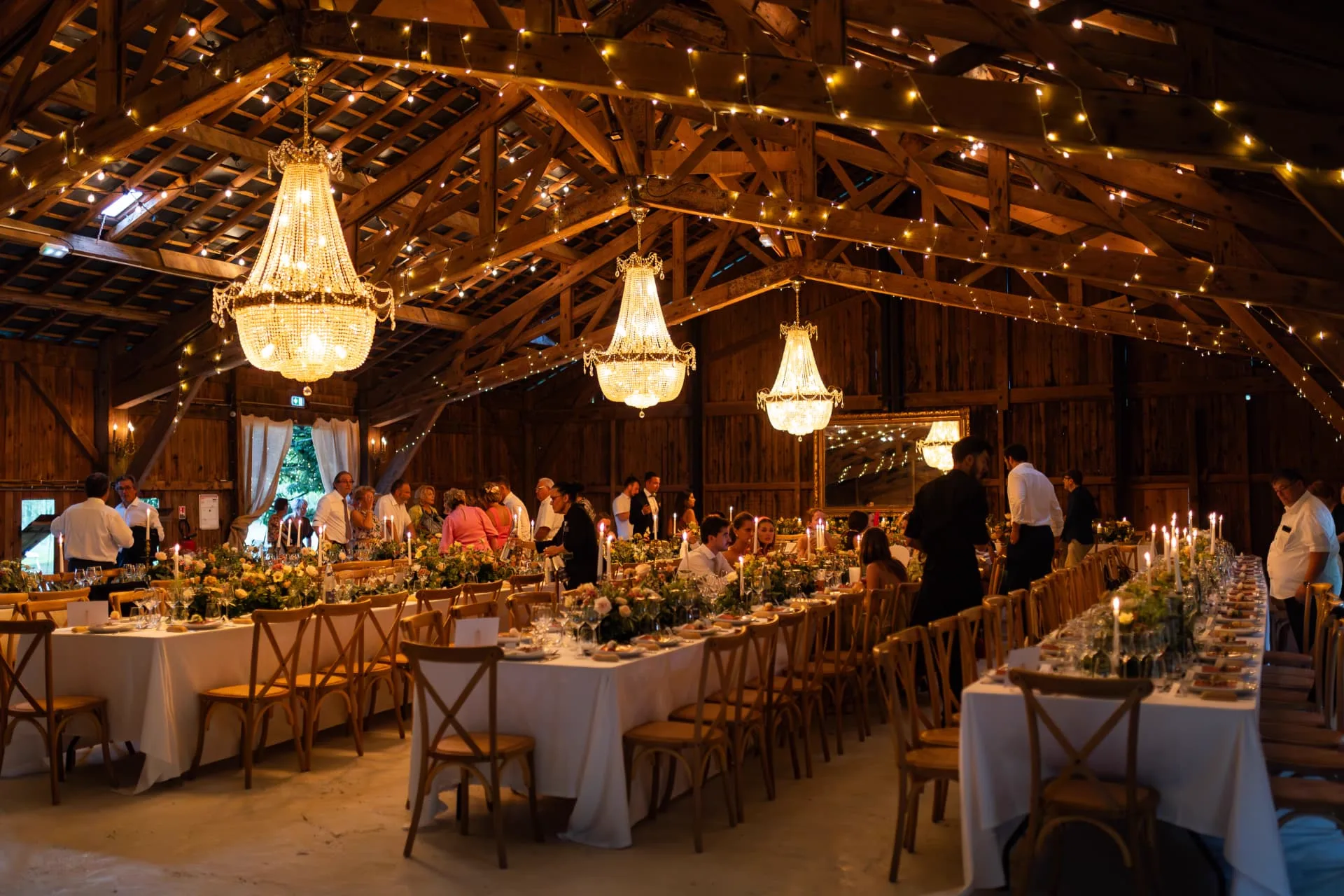 Salle de réception de mariage sous une charpente en bois, lustres et guirlandes lumineuses, longues tables fleuries dressées pour le dîner